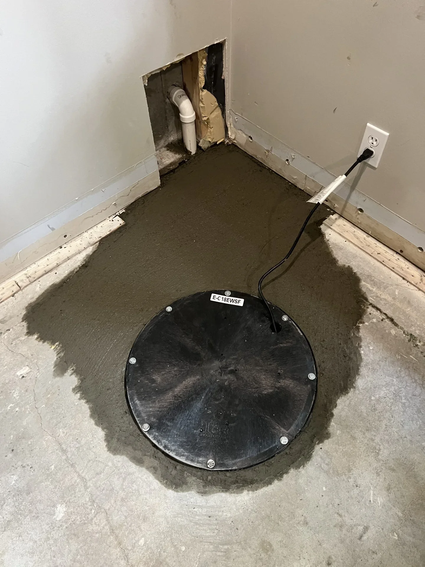 Black round sump basin cover set in fresh concrete with white PVC discharge pipe and power cord running to a wall outlet