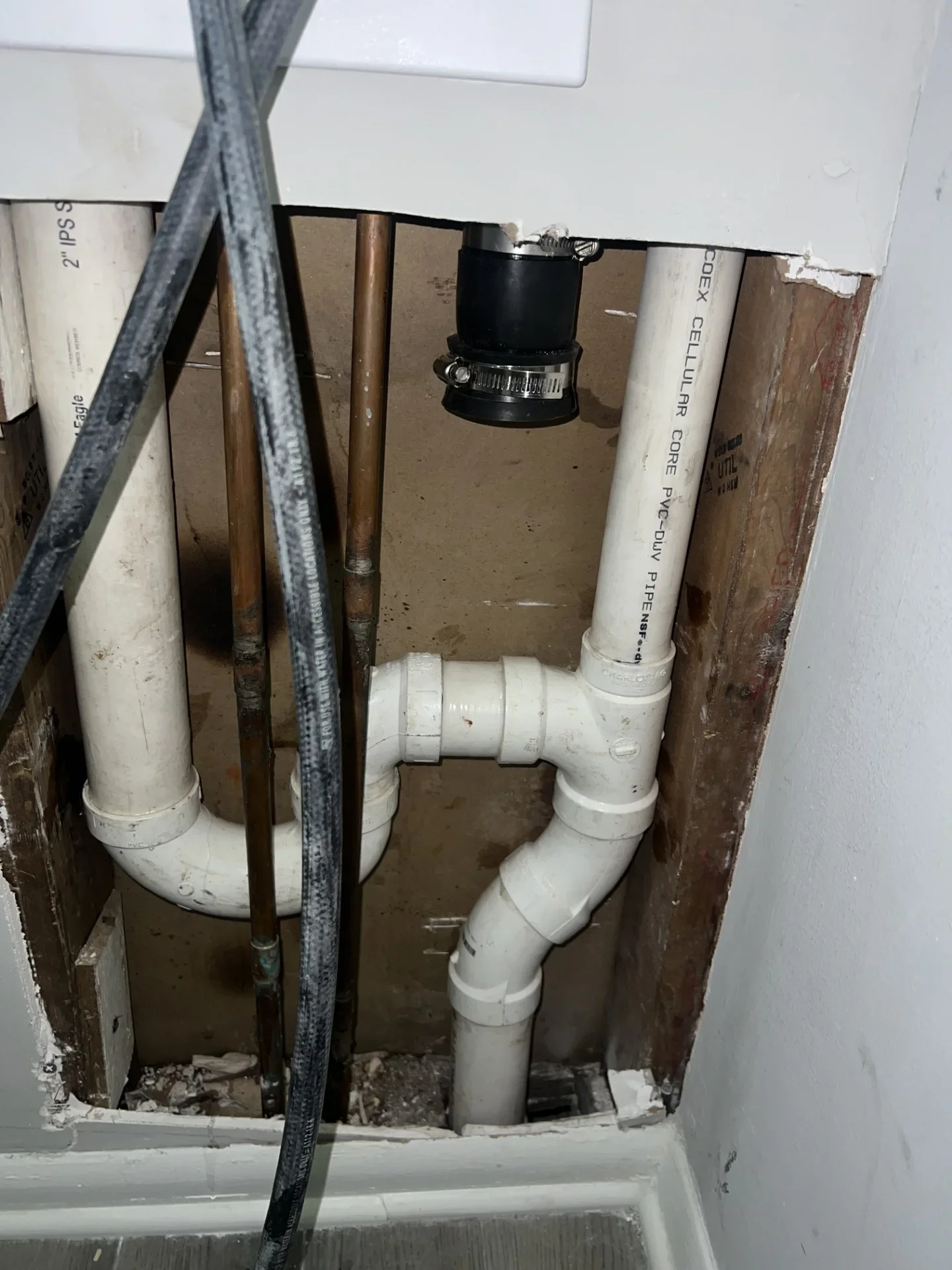Open wall cavity showing white PVC drain and vent piping, black rubber coupling with stainless clamps, and copper supply lines