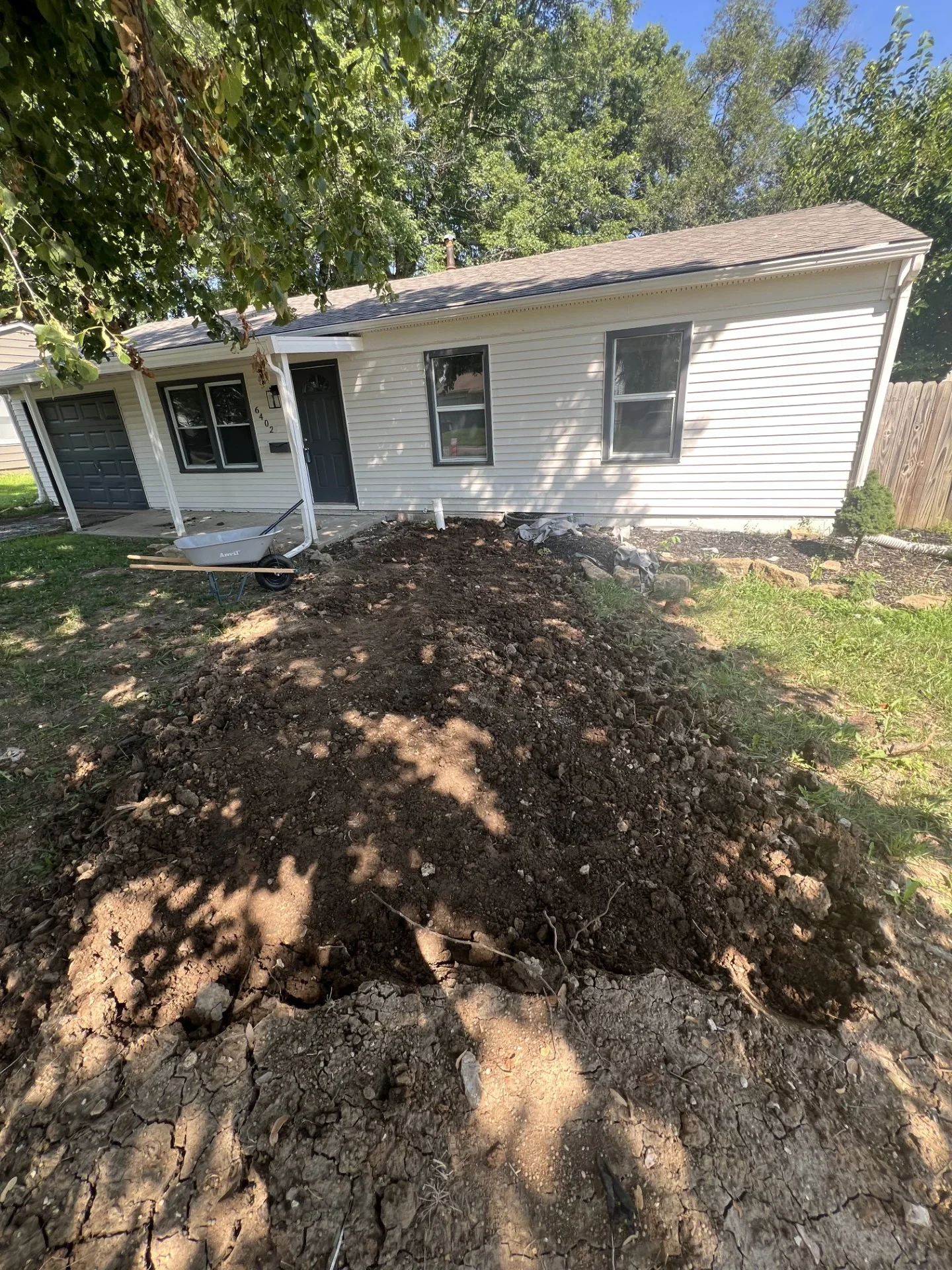Excavated dirt pile in front yard beside vinyl-sided house with wheelbarrow, showing trench work for underground line installation