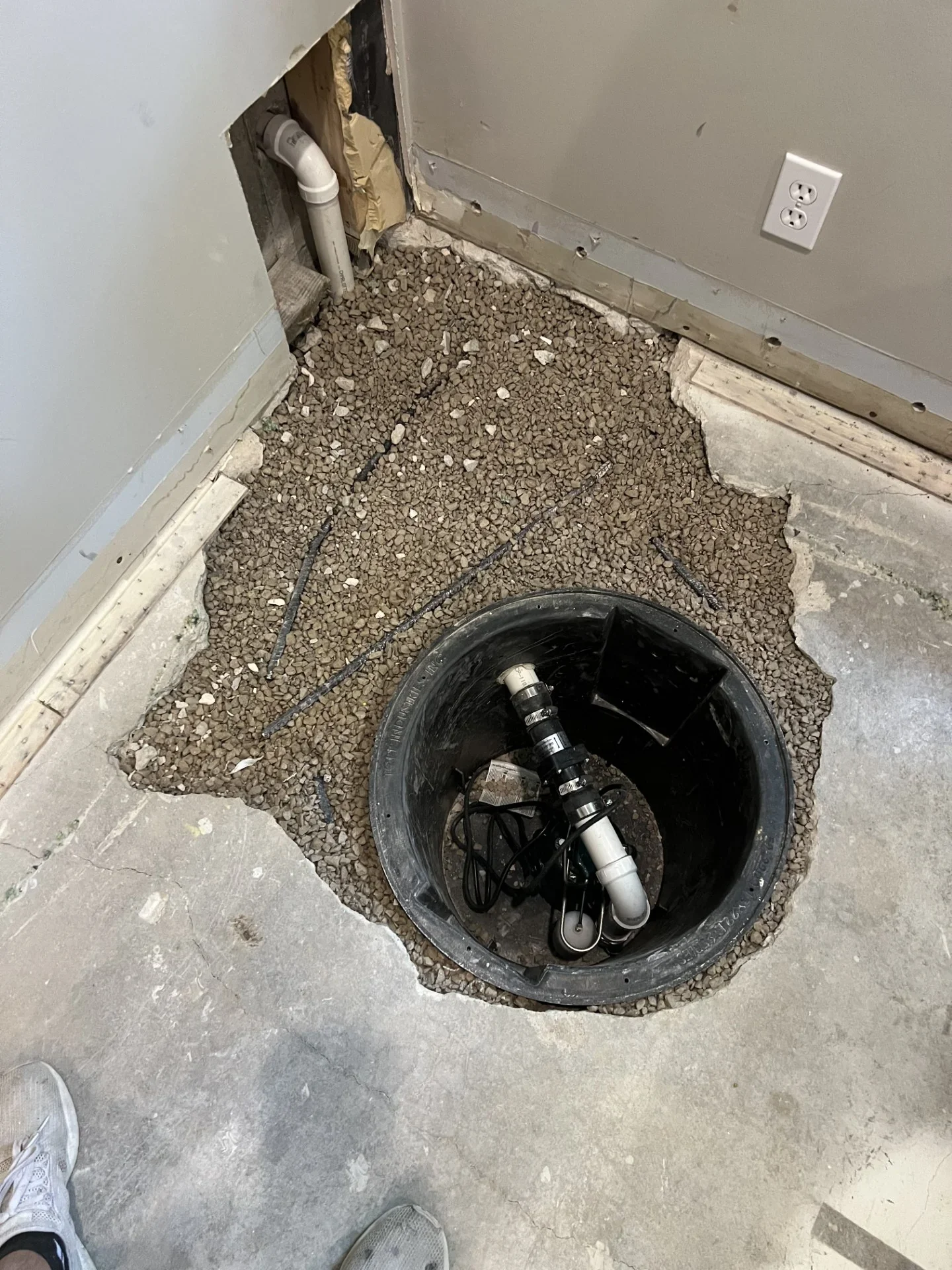 Black sump basin with submersible pump, white PVC discharge piping, and gravel fill set into a cut concrete basement floor