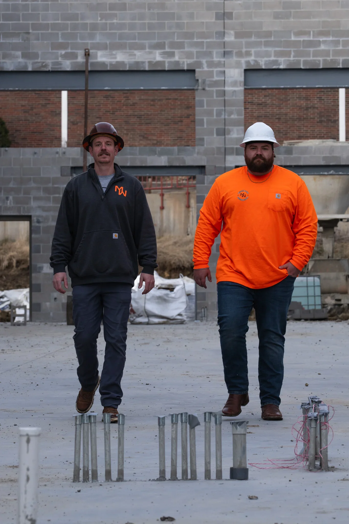 Two workers walking across a concrete slab past galvanized steel pipe sleeves and a capped PVC stub-out