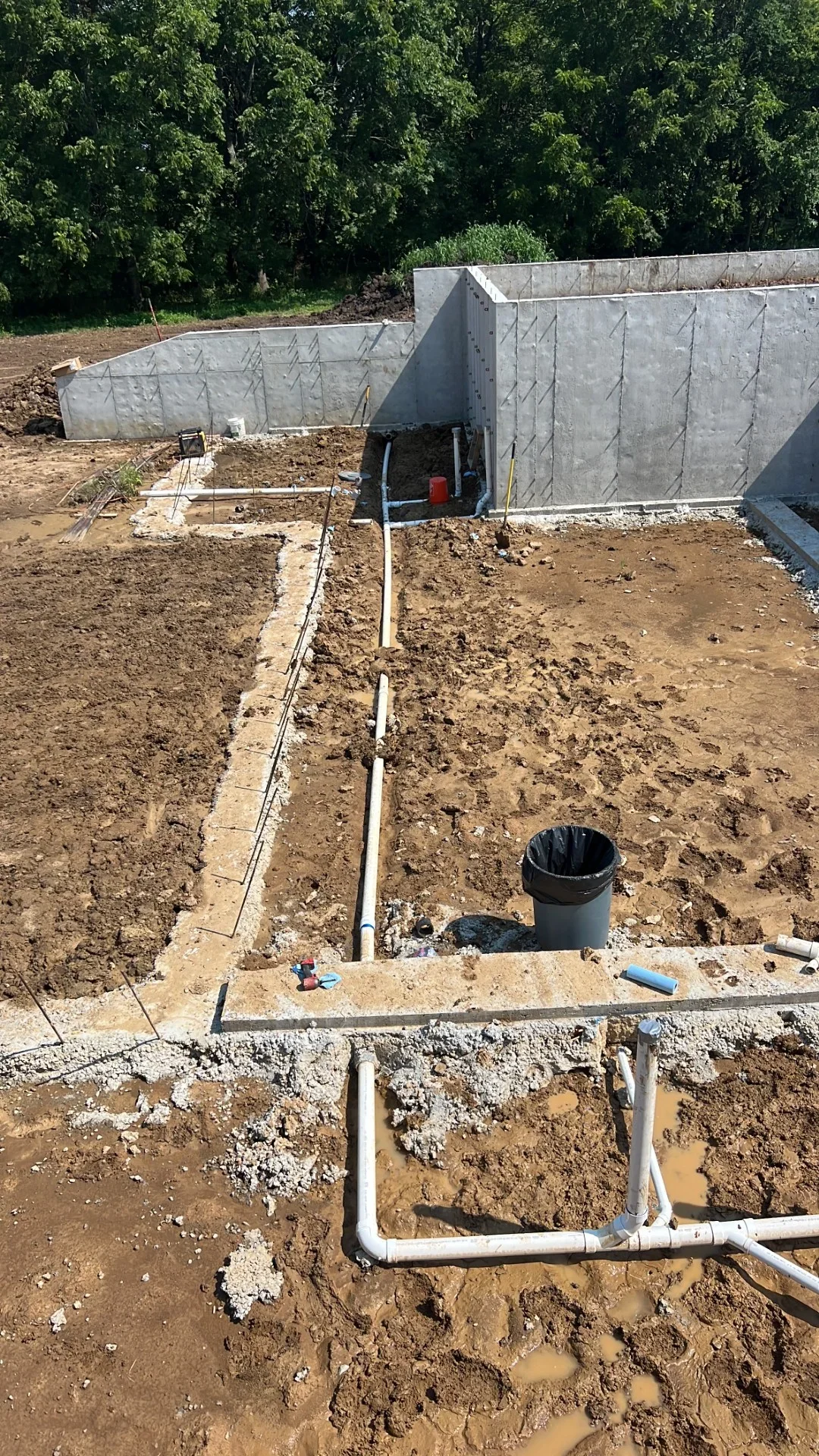 White PVC underground drain and supply lines with fittings in trenched soil beside poured concrete foundation walls and footings