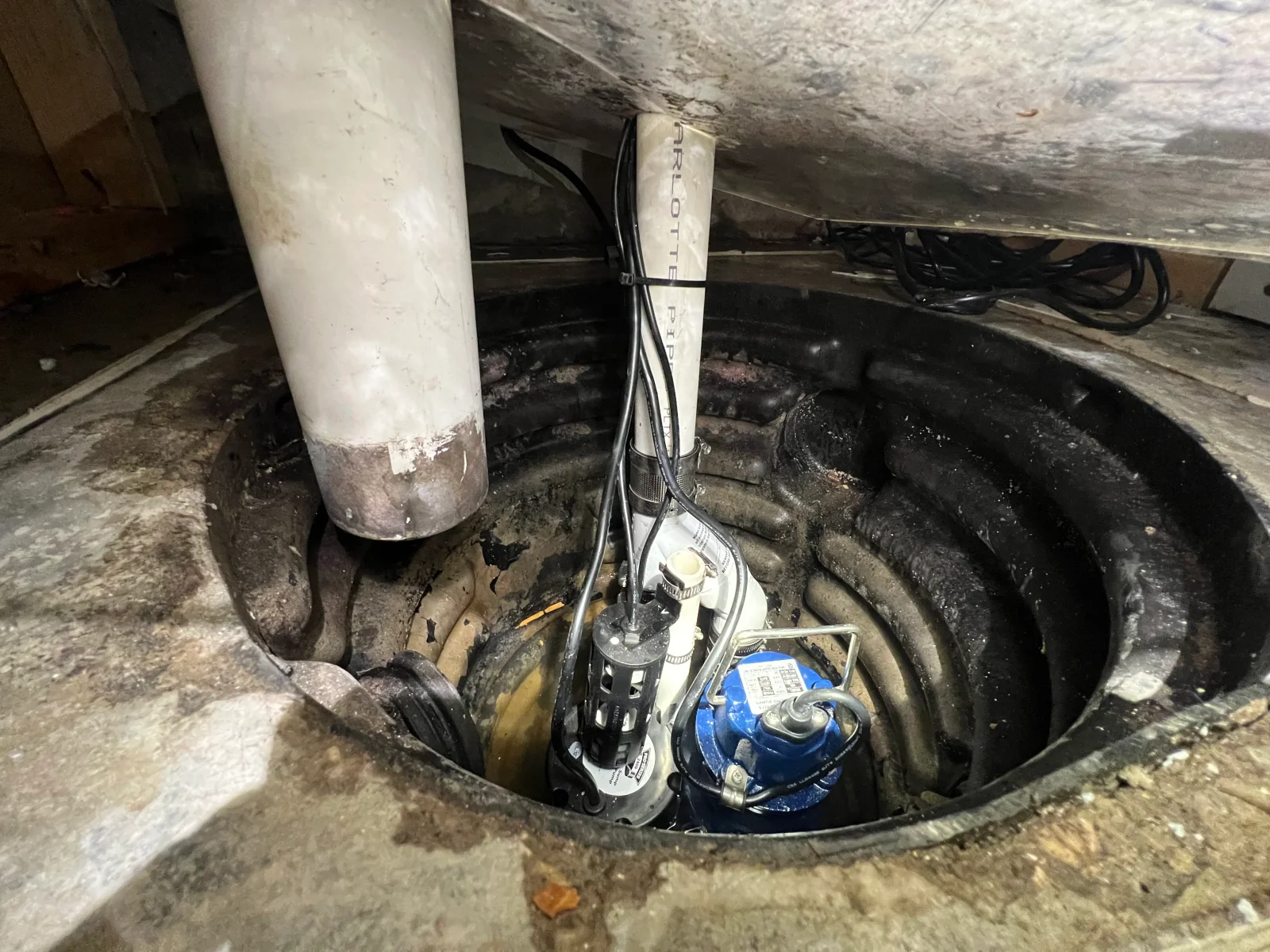 Blue submersible sump pump with white PVC discharge pipe and check valve installed in a black basin in a concrete crawl space floor