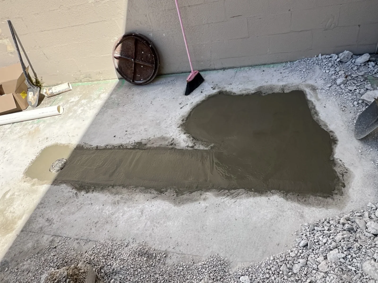Fresh gray concrete patch troweled over a slab beside a floor drain, with PVC pipe and hand tools nearby