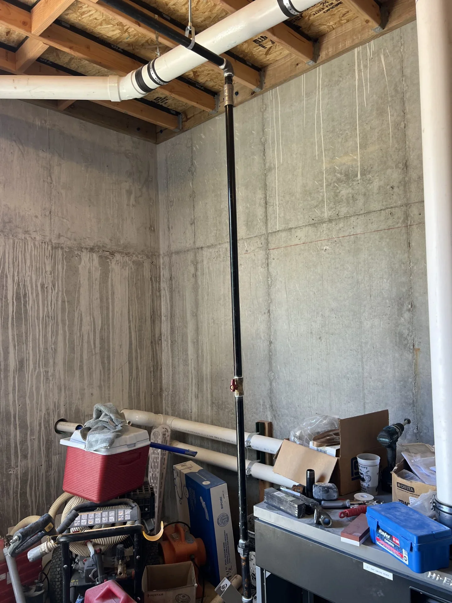 Black steel pipe riser with brass ball valve beside white PVC drain lines on exposed ceiling joists and concrete wall