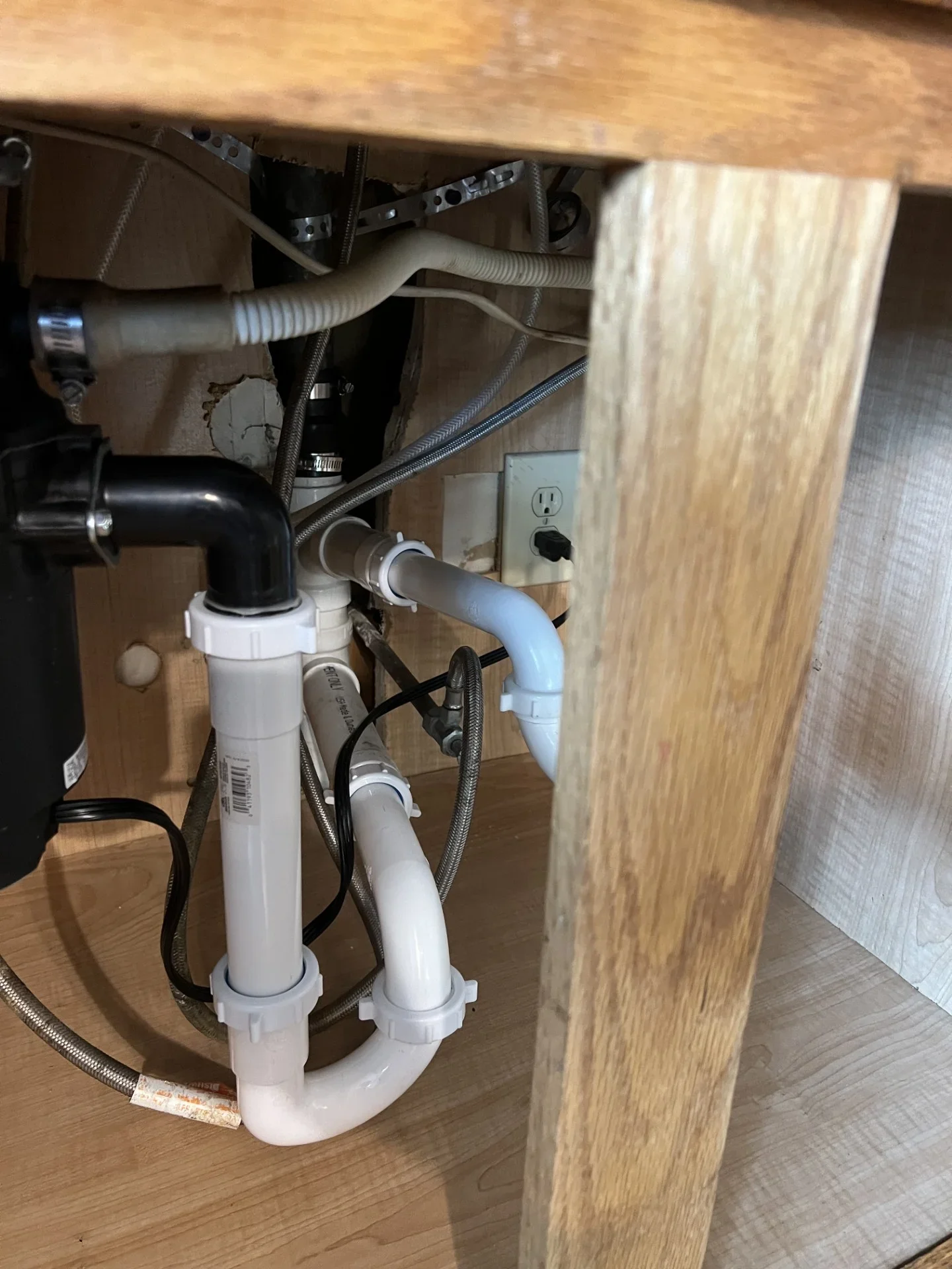 Under-sink PVC drain piping with P-trap, slip-joint nuts, black disposal elbow, and braided stainless steel supply lines in a wood cabinet