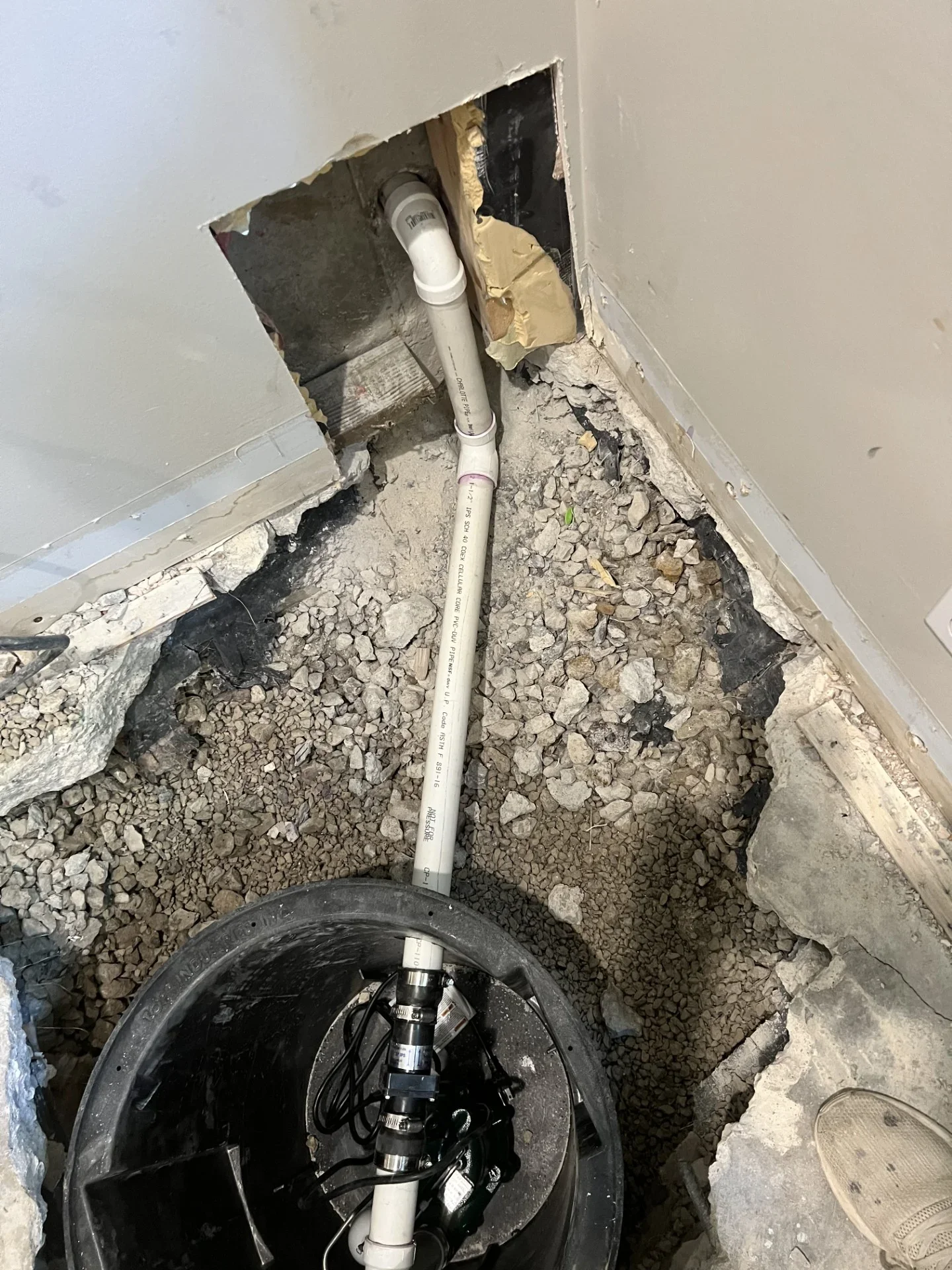 Black sump basin and pump with white PVC discharge pipe and elbow set in gravel-filled floor pit