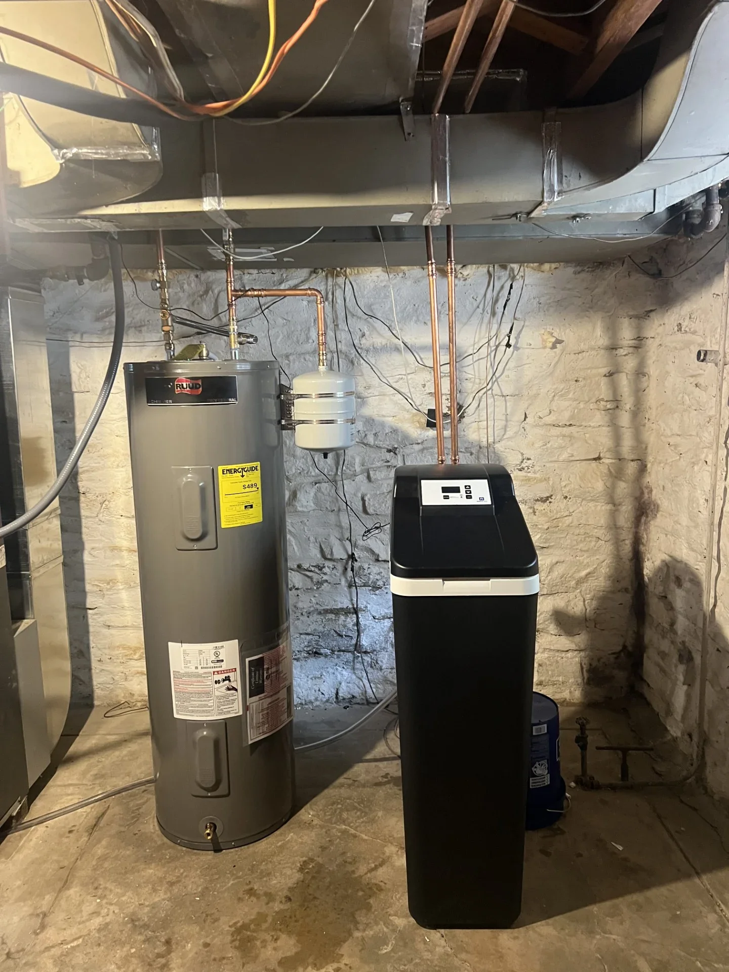 Black water softener with digital control panel beside a gray Ruud electric water heater and copper plumbing in a basement