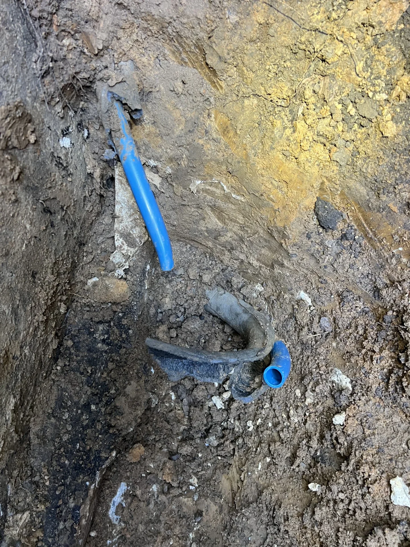Excavated trench showing blue PEX water line connected to a damaged gray underground pipe with blue elbow fitting
