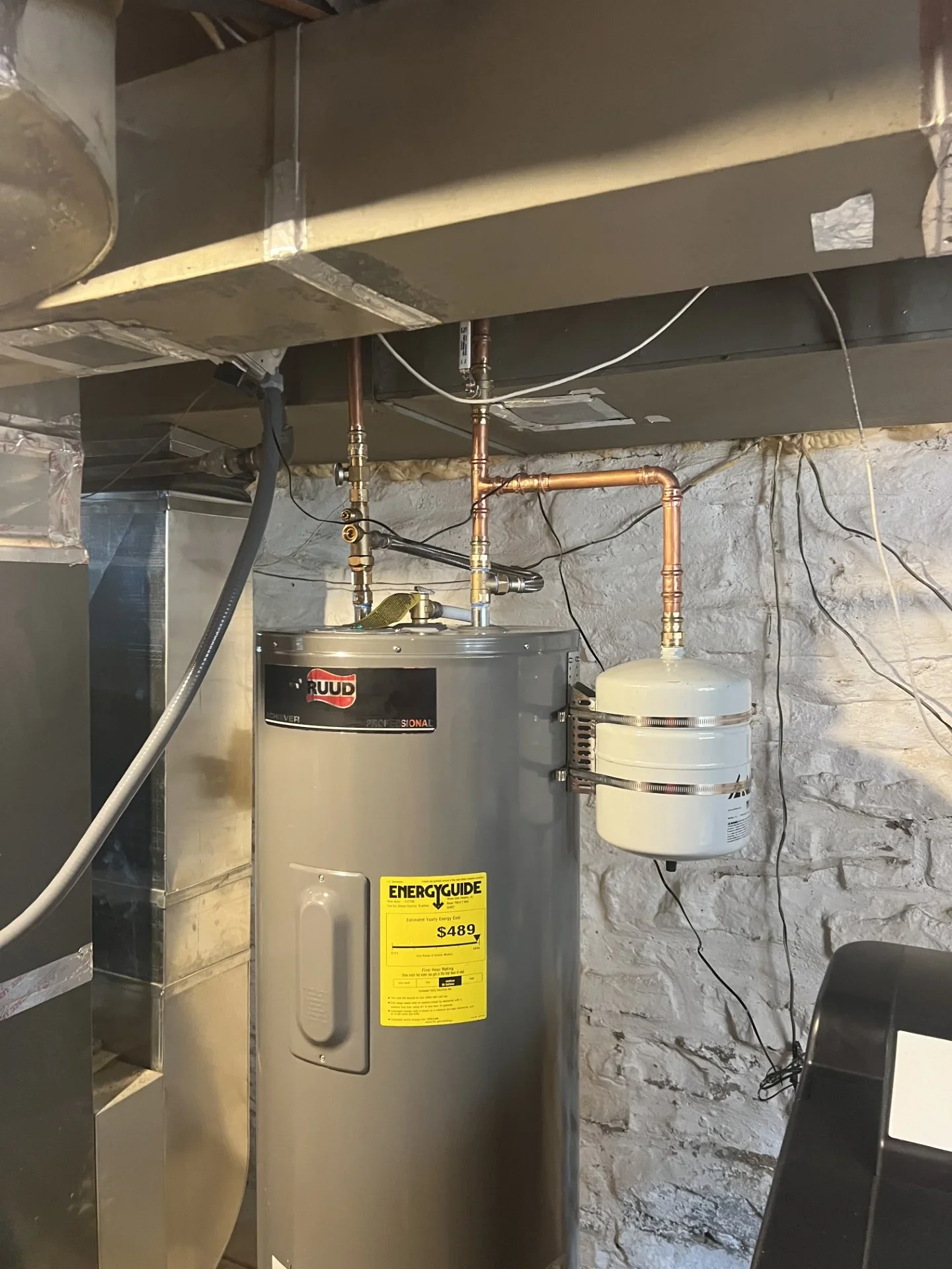 Gray electric water heater with copper piping, braided connectors, and white expansion tank strapped to a whitewashed stone wall