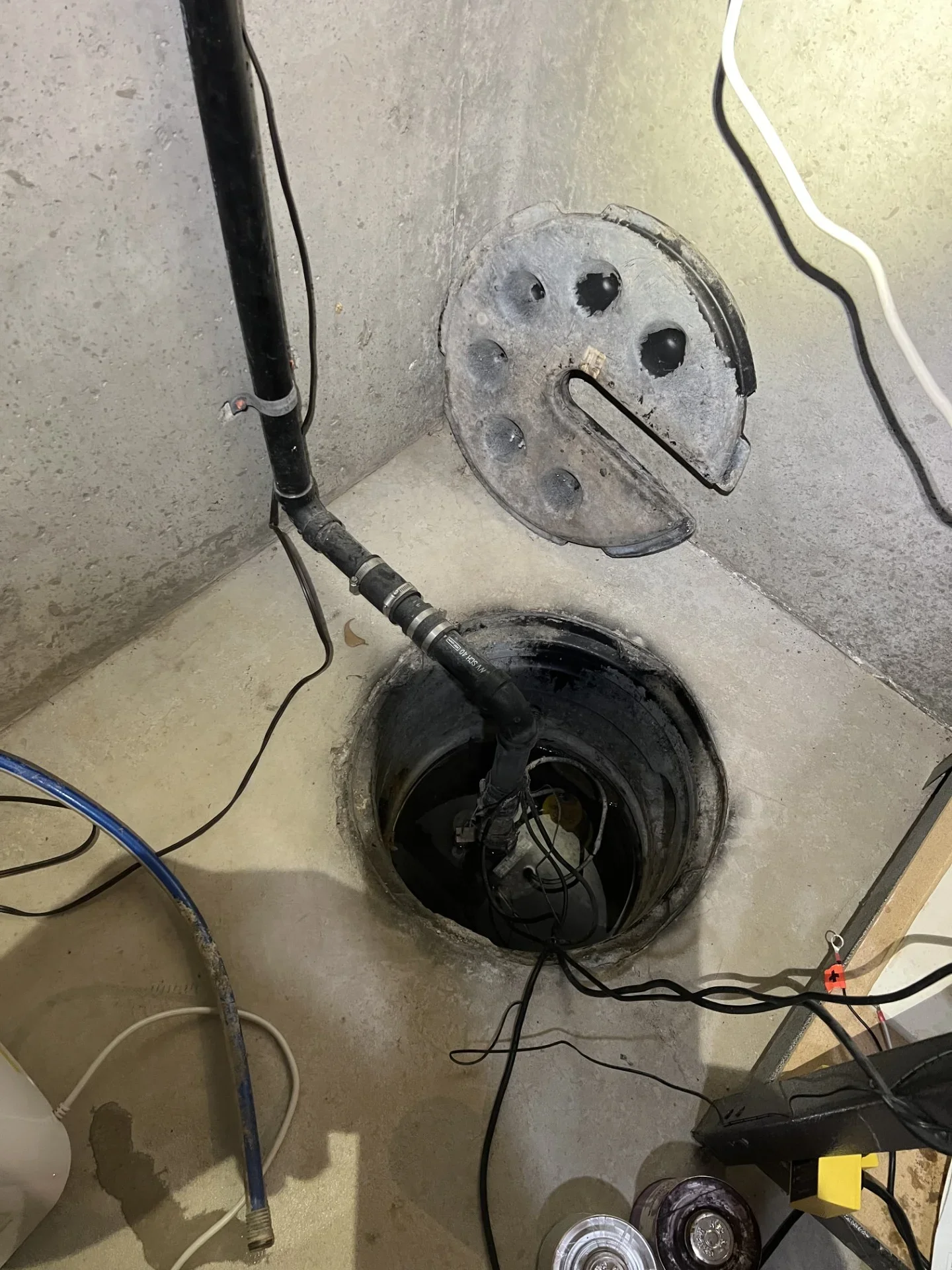 Submersible sump pump in a concrete basin with black PVC discharge pipe, hose-clamp couplings, and gray plastic basin cover