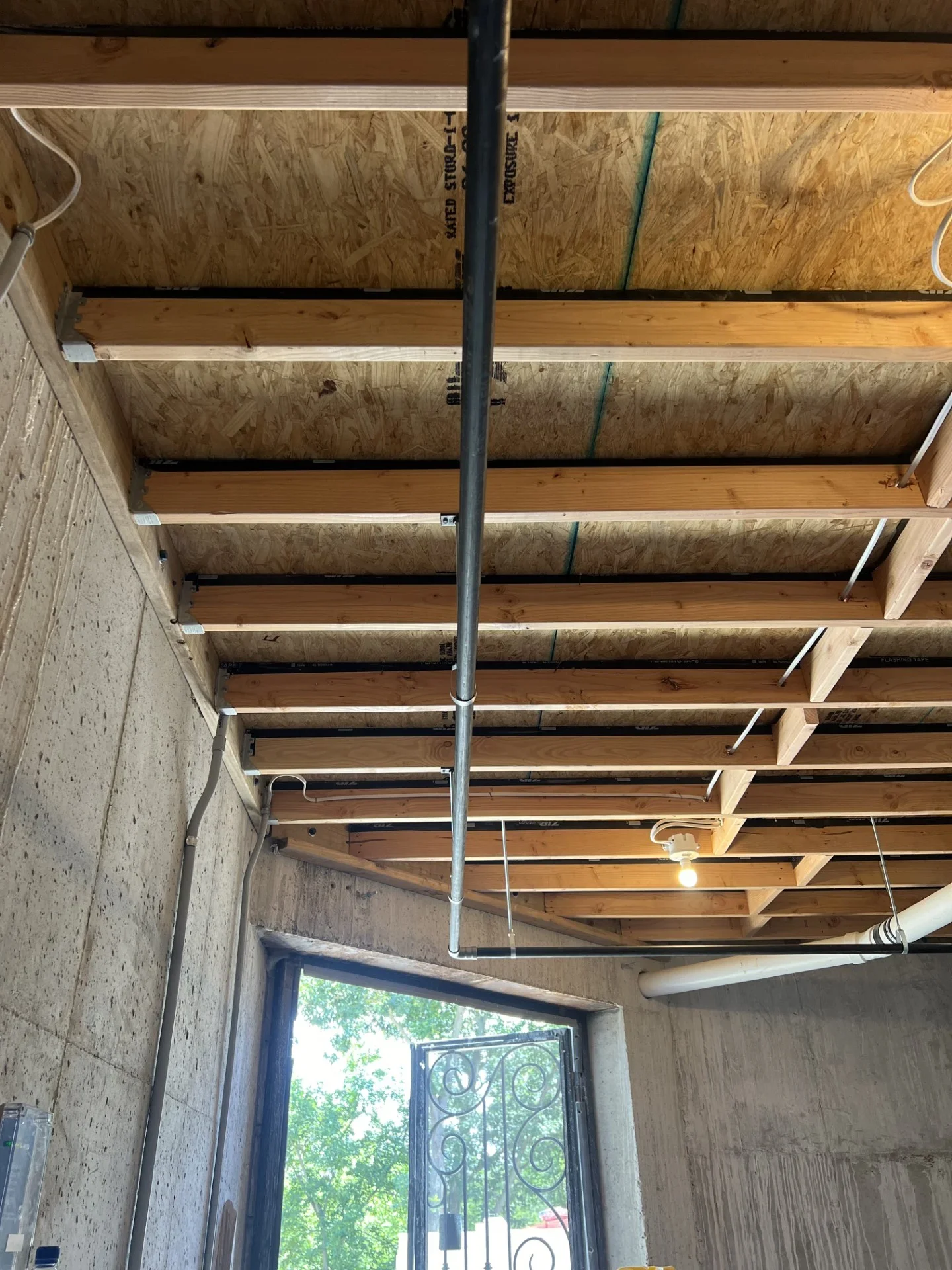 Exposed ceiling joists with OSB decking, galvanized steel pipe run, and PVC drain line in a concrete-walled room